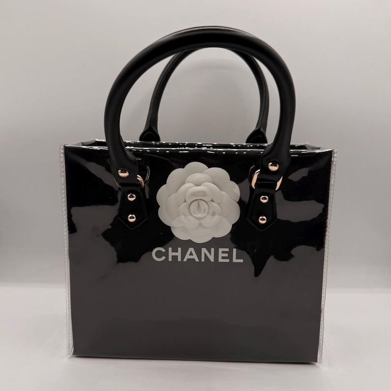 May include: A black glossy handbag with a structured rectangular shape, featuring two black top handles with gold-tone hardware. A large white camellia flower emblem is centered on the front, positioned above the white text CHANEL.