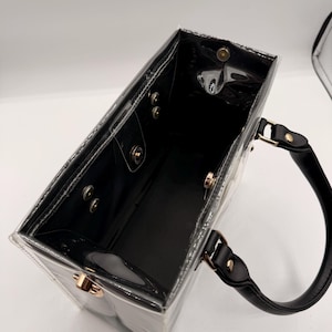 May include: A rectangular clear plastic tote bag with black trim and black handles. The interior features a black base and sides with gold-toned metal snap button closures along the top rim and side panels for secure fastening of the bag structure.