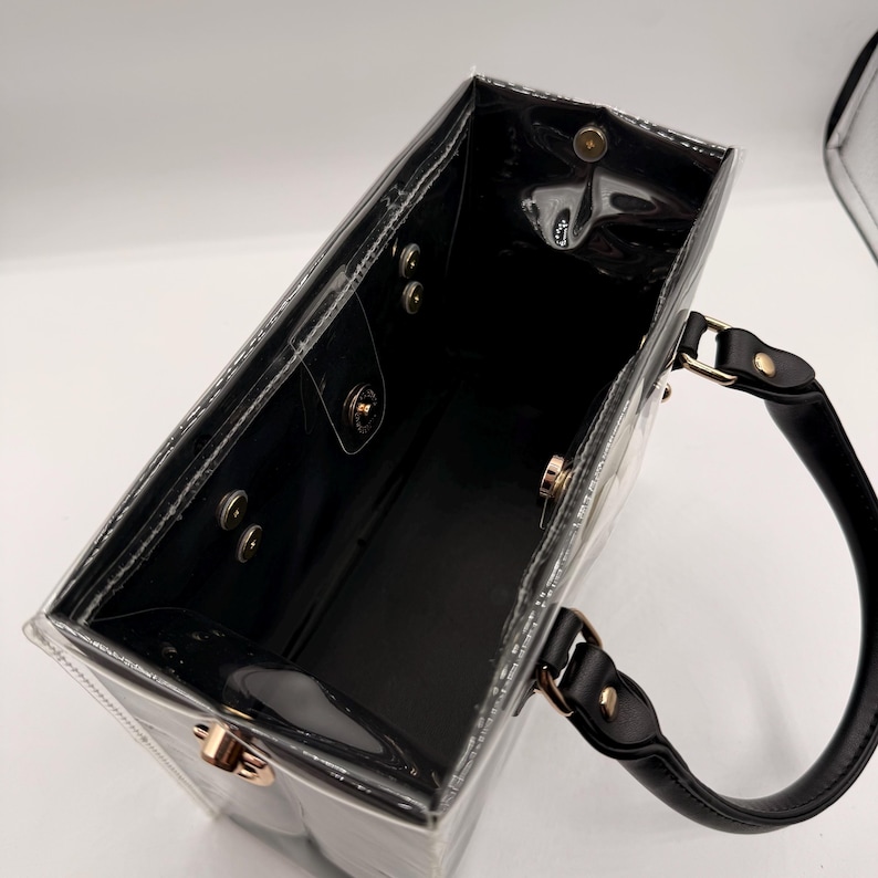 May include: A rectangular clear plastic tote bag with black trim and black handles. The interior features a black base and sides with gold-toned metal snap button closures along the top rim and side panels for secure fastening of the bag structure.