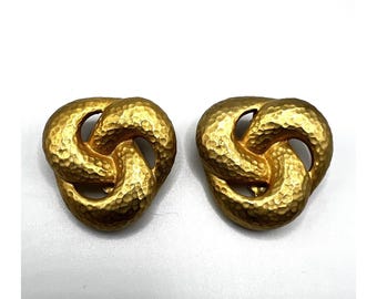 May include: A pair of gold-colored, textured knot-shaped earrings. The earrings have a hammered metal appearance and a three-dimensional design. The earrings are displayed on a white surface.