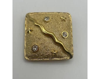 May include: A gold-toned square brooch with a textured surface. A wavy gold line bisects the square. The brooch features three clear, round gemstones and three star-shaped accents. The brooch is a vintage accessory.