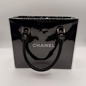May include: A black rectangular handbag with a glossy finish and clear side panels. The front features the word CHANEL in white lettering. It includes two black handles attached with gold-toned hardware, providing a structured look for this accessory.
