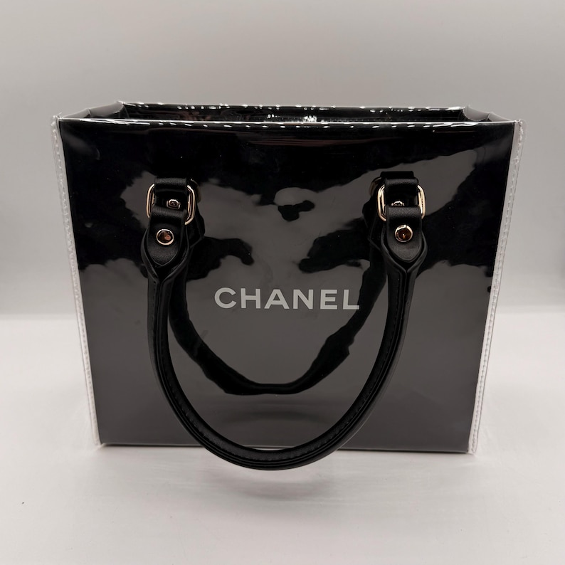 May include: A black rectangular handbag with a glossy finish and clear side panels. The front features the word CHANEL in white lettering. It includes two black handles attached with gold-toned hardware, providing a structured look for this accessory.