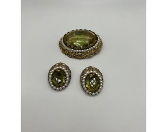 May include: A vintage jewelry set featuring a large oval brooch and matching earrings. The set has a gold-tone metal setting with a central olive-green stone, surrounded by small faux pearls. The brooch and earrings are on a white background.