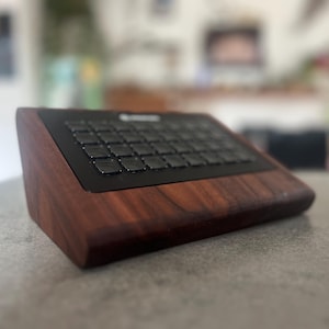 以下が含まれることがあります： 濃い木材仕上げと黒いキーボードを備えた木製キーボード。 キーボードは角度がついており、洗練されたモダンなデザインです。 キーは黒と正方形です。 キーボードは灰色の表面に置かれています。