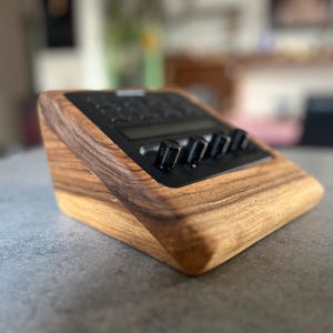 以下が含まれることがあります： 黒いコントロールパネルとノブが付いた木製の電子デバイス。デバイスは傾斜したデザインで、木材で作られており、木目が見えます。ノブは黒色で、オーディオ機器のようです。