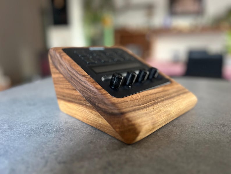 以下が含まれることがあります： 黒いコントロールパネルとノブが付いた木製の電子デバイス。デバイスは傾斜したデザインで、木材で作られており、木目が見えます。ノブは黒色で、オーディオ機器のようです。