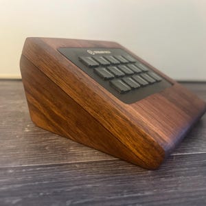 May include: A dark brown wooden housing for a Stream Deck device, featuring a slanted wedge shape with a smooth, polished grain finish. The black control panel is recessed into the top surface, displaying a grid of square buttons for input.