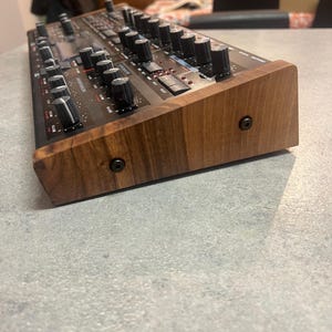 Op de afbeelding: Een donkergrijze synthesizer met een houten zijpaneel. De synthesizer heeft veel knoppen en draaiknoppen, en een klein digitaal display.