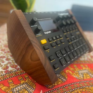 Könnte beinhalten: Ein schwarzer Elektron Digitakt Drumcomputer und Sampler mit zentralem Display, zahlreichen Drehreglern und einem Raster aus hintergrundbeleuchteten Tasten, untergebracht in einem dunklen Holzständer für eine angewinkelte Positionierung.