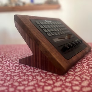 May include: A dark brown wooden stand holding a black electronic device with a grid of square buttons, a small rectangular display screen, and a row of five circular control knobs positioned along the bottom edge of the slanted interface panel.
