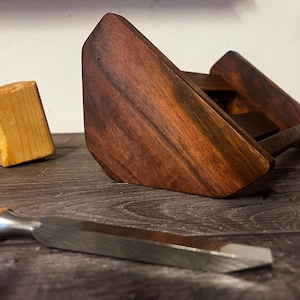 May include: A dark brown wooden stand with a tiered design, featuring a smooth, polished finish and visible wood grain. Beside it lies a light-colored wooden mallet and a metal chisel with a wooden handle, showcasing a natural wood aesthetic.