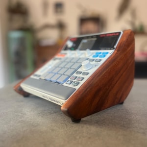 Könnte beinhalten: Ein Musik-Sampler-Gerät mit weißem Bedienfeld, grauen Pads, blauen Tasten und einem schwarzen Display, montiert in einem maßgefertigten, angewinkelten Holzständer mit einer satten, dunkelbraunen Holzmaserung und kleinen Standfüßen.