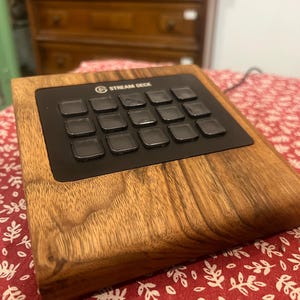 Könnte beinhalten: Ein Stream Deck mit Holzsockel und schwarzem Bedienfeld. Das Stream Deck hat 15 schwarze quadratische Tasten und das Stream Deck-Logo. Der Holzsockel hat eine natürliche Holzmaserung.