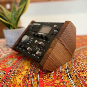 Op de afbeelding: Een zwarte elektronische muzieksynthesizer met houten zijpanelen. Het apparaat heeft talrijke knoppen, toetsen en een klein scherm. Het staat op een kleurrijk patroon, met een plant in een pot op de achtergrond.