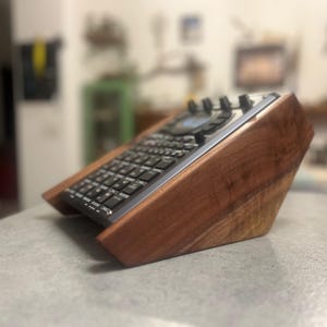 Puede incluir: Un dispositivo muestreador de música electrónica gris oscuro con una cuadrícula de botones y una pantalla pequeña, montado en posición inclinada dentro de un soporte de madera marrón oscuro pulido a medida con un acabado de veta natural.