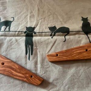 Op de afbeelding: Twee houten deurvastzetters met een natuurlijk houtnerfpatroon, elk met twee gaten. De deurvastzetters liggen op een beige stof met een herhalend patroon van zwarte katten silhouetten.