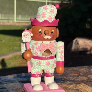 Pode incluir: Um boneco quebra-nozes com tema natalino com rosto castanho, sardas e bigode branco. A boneca usa uma roupa com estampa floral verde menta e rosa, um la&ccedil;o rosa e uma coroa. Ele segura um enfeite de Papai Noel e fica em uma base rosa com "Merry Christmas" escrito.