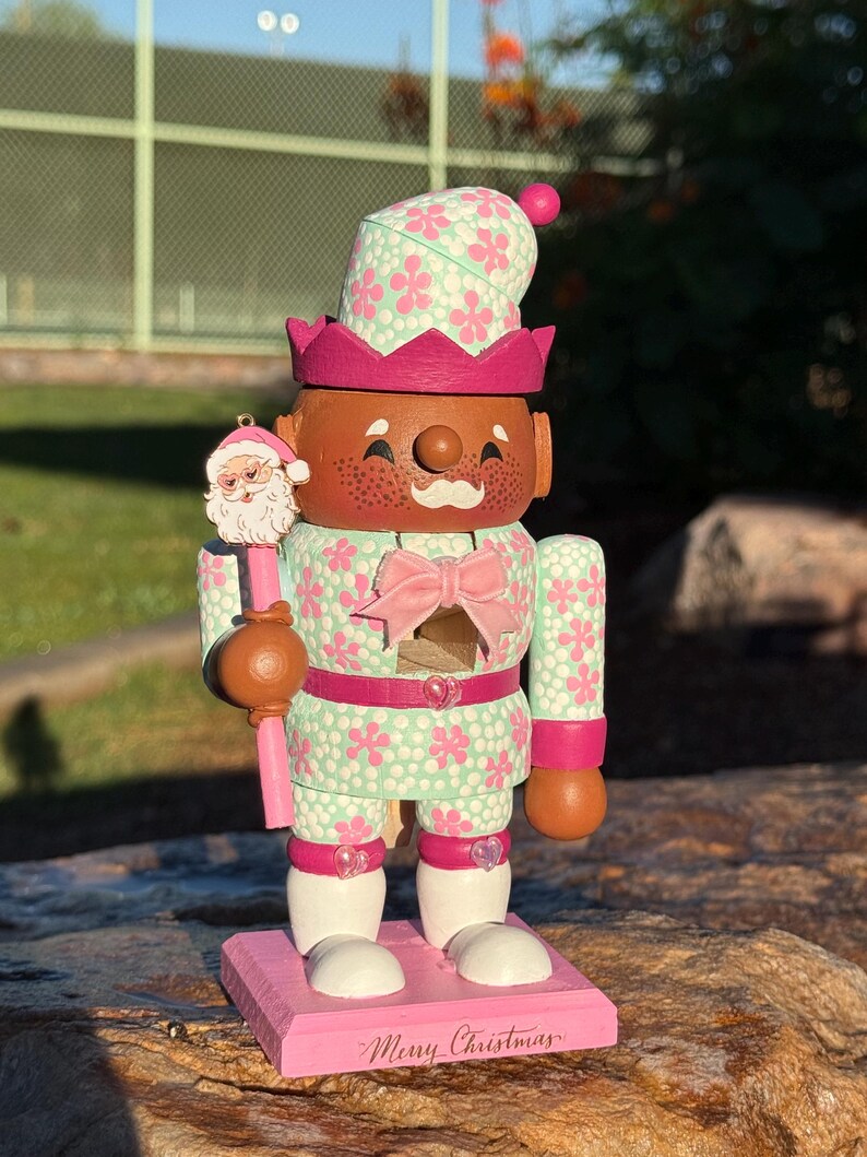 Pode incluir: Um boneco quebra-nozes com tema natalino com rosto castanho, sardas e bigode branco. A boneca usa uma roupa com estampa floral verde menta e rosa, um la&ccedil;o rosa e uma coroa. Ele segura um enfeite de Papai Noel e fica em uma base rosa com "Merry Christmas" escrito.