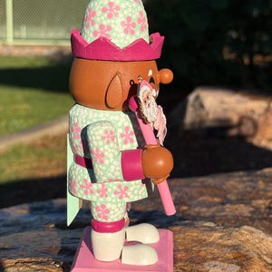 Pode incluir: Uma figura de quebra-nozes de madeira com um rosto castanho, vestindo uma roupa e chap&eacute;u com estampa floral verde menta e rosa. O quebra-nozes segura uma bengala rosa com um enfeite de Papai Noel. A base &eacute; rosa.