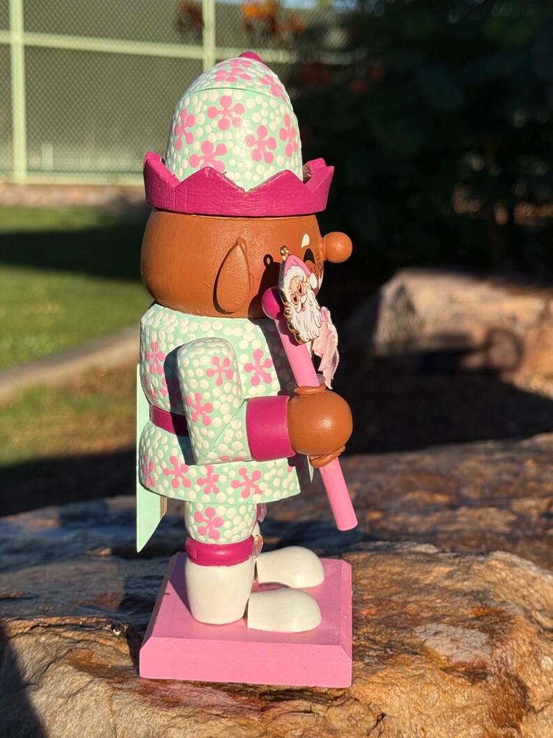 Pode incluir: Uma figura de quebra-nozes de madeira com um rosto castanho, vestindo uma roupa e chap&eacute;u com estampa floral verde menta e rosa. O quebra-nozes segura uma bengala rosa com um enfeite de Papai Noel. A base &eacute; rosa.