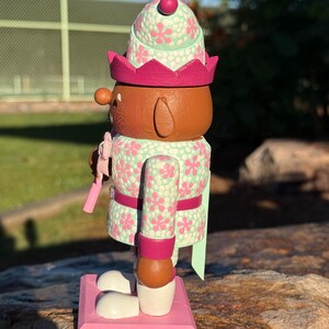 Pode incluir: Figura decorativa de quebra-nozes de madeira. O quebra-nozes &eacute; pintado com um padr&atilde;o floral verde claro e rosa. Possui um rosto castanho, uma coroa rosa e uma base rosa. A figura est&aacute; sobre uma base rosa.