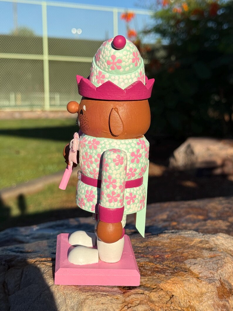 Pode incluir: Figura decorativa de quebra-nozes de madeira. O quebra-nozes &eacute; pintado com um padr&atilde;o floral verde claro e rosa. Possui um rosto castanho, uma coroa rosa e uma base rosa. A figura est&aacute; sobre uma base rosa.