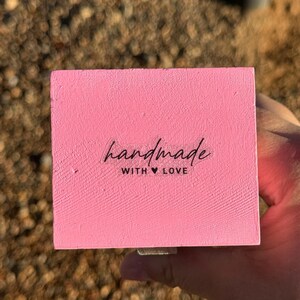 Pode incluir: Um bloco de madeira retangular rosa com as palavras "handmade with love" em escrita preta. O bloco &eacute; segurado em uma m&atilde;o, com um fundo desfocado marrom e verde.