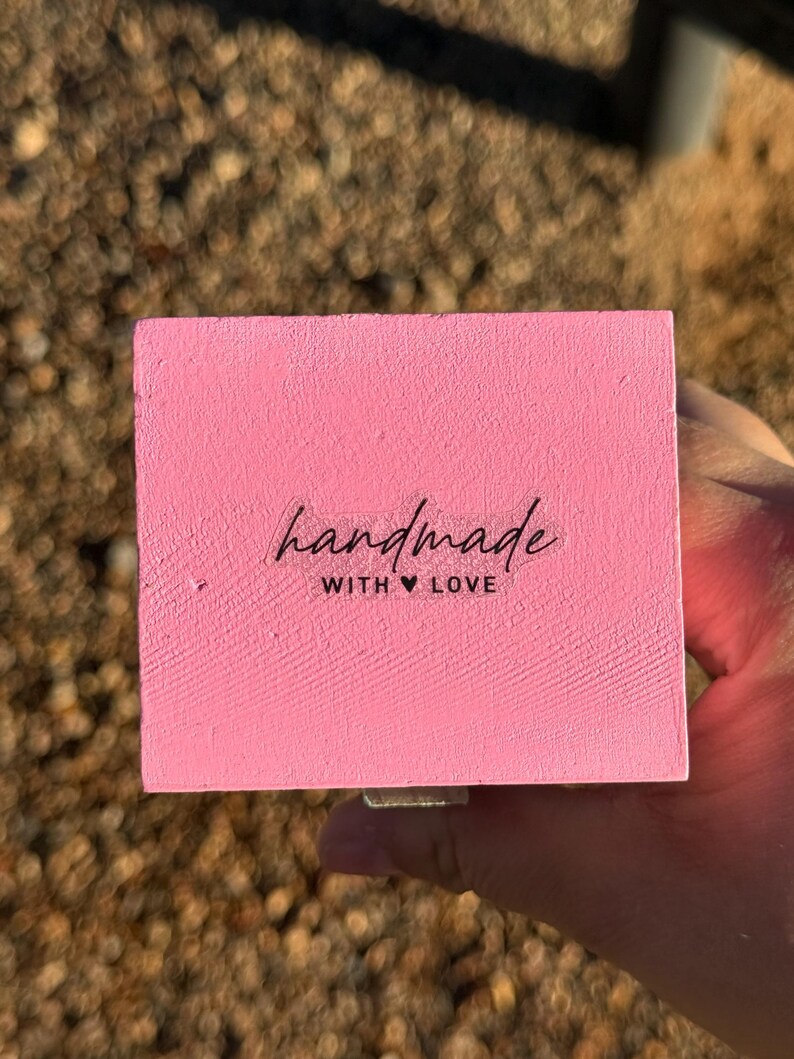 Pode incluir: Um bloco de madeira retangular rosa com as palavras "handmade with love" em escrita preta. O bloco &eacute; segurado em uma m&atilde;o, com um fundo desfocado marrom e verde.