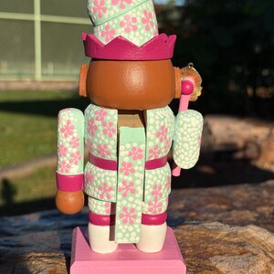 Pode incluir: Figura de quebra-nozes de madeira pintada &agrave; m&atilde;o. O quebra-nozes tem um rosto castanho, uma roupa com estampa floral verde menta e rosa e uma base rosa. O chap&eacute;u e a roupa s&atilde;o decorados com flores rosa. O quebra-nozes est&aacute; sobre uma base rosa.