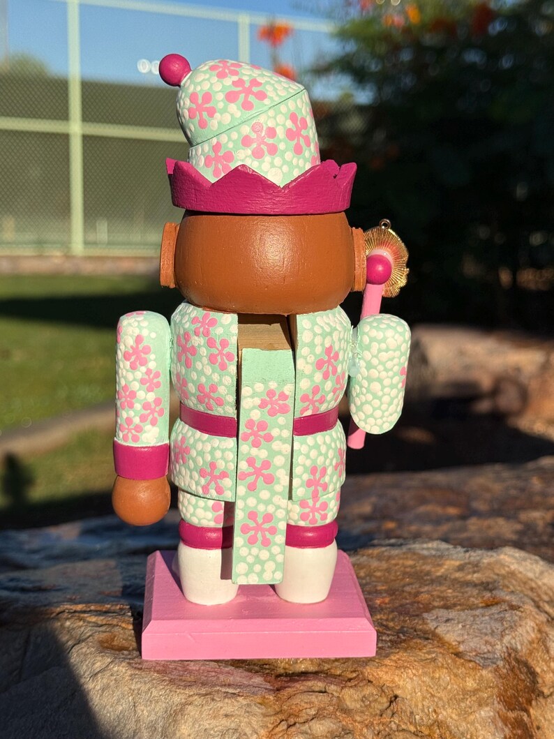 Pode incluir: Figura de quebra-nozes de madeira pintada &agrave; m&atilde;o. O quebra-nozes tem um rosto castanho, uma roupa com estampa floral verde menta e rosa e uma base rosa. O chap&eacute;u e a roupa s&atilde;o decorados com flores rosa. O quebra-nozes est&aacute; sobre uma base rosa.