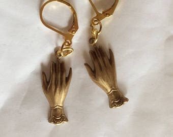 May include: A pair of gold earrings featuring a hand design. The earrings have a leverback closure.