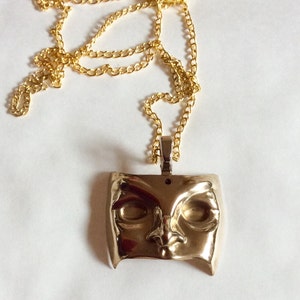 Op de afbeelding: Een gouden ketting met een gouden hanger in de vorm van een gestileerd menselijk gezichtsmasker.