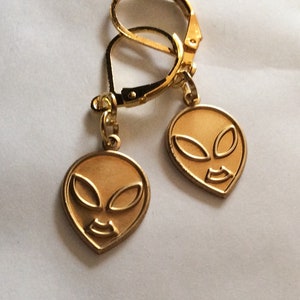 Peut inclure: Boucles d'oreilles dorées à fermoir à levier avec un pendentif en forme de visage d'extraterrestre. Le visage extraterrestre a de grands yeux ovales et une petite bouche incurvée.