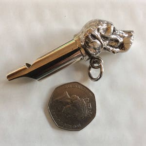 May include: Silver dog whistle with a detailed dog head design. The whistle is about the size of a British 20 pence coin.
