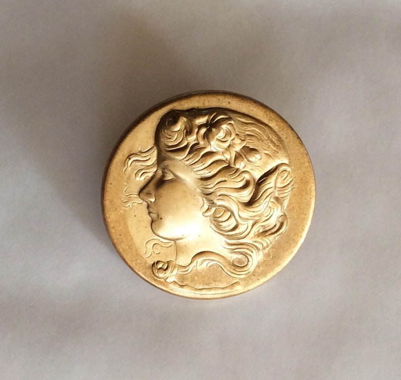 May include: A gold-toned metal compact with a raised design of a woman's profile. The woman has long, flowing hair and is wearing a floral crown.