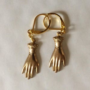 Könnte beinhalten: Ein Paar goldene Ohrringe mit einem Handdesign. Die Ohrringe sind aus Metall und haben ein schlichtes, elegantes Design.