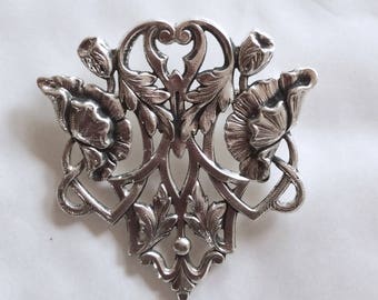Peut inclure: Une broche en argent avec un design délicat représentant des coquelicots et des feuilles stylisés. La broche est finement travaillée avec un motif de filigrane et a un centre en forme de cœur.