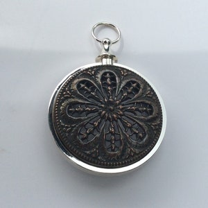 Könnte beinhalten: Ein silberner Anhänger mit einem runden, verzierten Kupfer-Mittelstück, das ein detailliertes Blumenmuster aufweist. Der Anhänger hat oben eine Schlaufe zum Anbringen einer Kette.