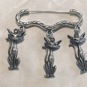 Peut inclure: Broche argentée avec trois breloques de chat suspendues. La broche est de style épingle de sûreté avec un design tressé. Les chats sont en position assise, la queue enroulée.