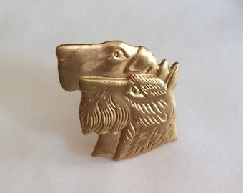 Op de afbeelding: Gouden metalen pin met twee hondenkoppen, de ene bovenop de andere. De kop van de bovenste hond is naar links gericht, terwijl de kop van de onderste hond naar rechts is gericht.