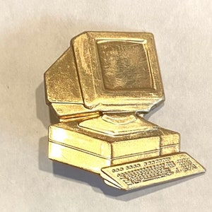 May include: A gold-coloured metal pin shaped like a vintage computer. The pin has a monitor, a keyboard, and a CPU unit.