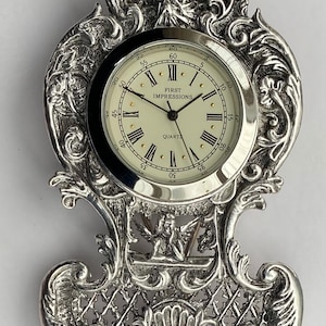 May include: An ornate silver-toned clock with a white face and black Roman numeral hour markers. The clock is on a decorative stand with a filigree design. The clock face reads "First Impressions Quartz".