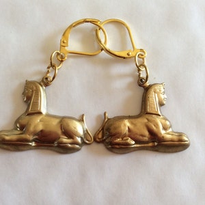 May include: A pair of gold-tone keyrings featuring a sculpted sphinx design. Each keyring has a split ring and a chain.