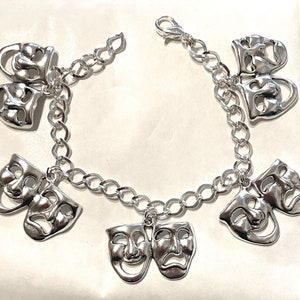 Op de afbeelding: Zilverkleurige bedelarmband met meerdere paren komedie- en tragediemaskers. De armband heeft een schakelontwerp met een karabijnsluiting. De maskers zijn gedetailleerd met gezichtskenmerken en hangen vrij aan de ketting.