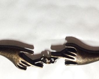 Op de afbeelding: Een gouden ketting met twee handen die als hanger aan elkaar vastzitten. De handen zijn van metaal en hebben een vintage uitstraling.