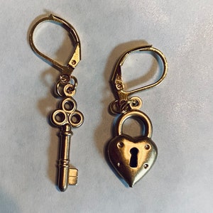 Puede incluir: Un par de pendientes de oro, uno con forma de llave y el otro con forma de candado con un diseño en forma de corazón.