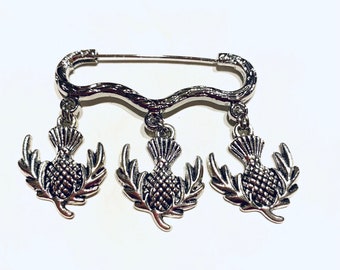 Puede incluir: Broche plateada con tres colgantes de cardo. La broche tiene forma de línea curva y cierre de seguridad.