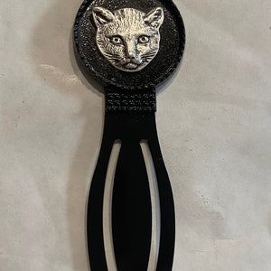 May include: Black metal bookmark with a silver cat head design. The cat is wearing a crown.