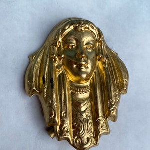 Peut inclure: Une broche en métal doré représentant un relief détaillé d'un visage de femme. La femme porte une coiffe et une robe flottante aux motifs complexes.